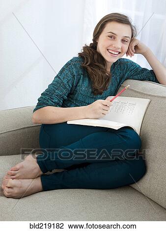 Stock Image - Smiling teenage girl doing homework on sofa. Fotosearch