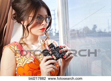 Stock Image - Woman holding binoculars at window. Fotosearch