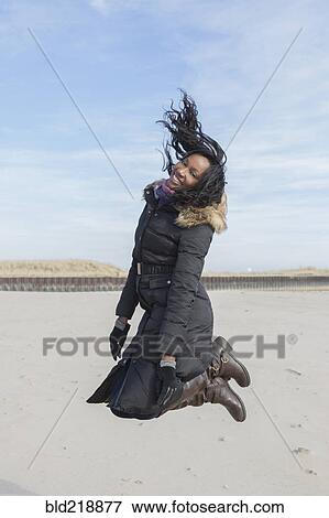 Stock Photo - Woman in coat jumping for joy on beach. Fotosearch