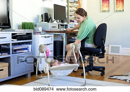 Picture - Caucasian mother working in home office with baby next to her. Fotosearch