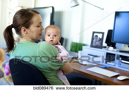 Stock Photograph - Caucasian mother working in home office and holding baby. Fotosearch