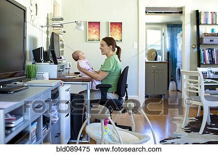 Stock Photography - Caucasian mother working in home office and holding baby. Fotosearch
