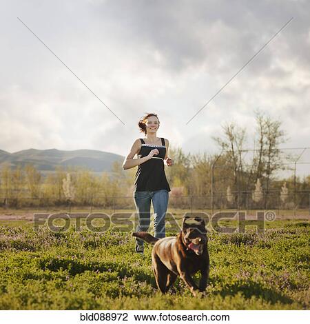 Stock Image - Caucasian woman running in field with dog. Fotosearch