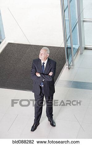 Stock Image - Hispanic businessman standing in lobby with arms crossed. Fotosearch