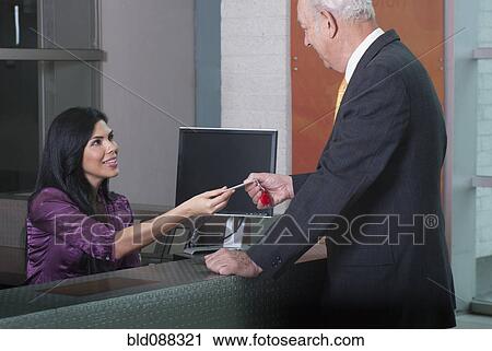 Stock Image - Hispanic receptionist handing card to businessman. Fotosearch
