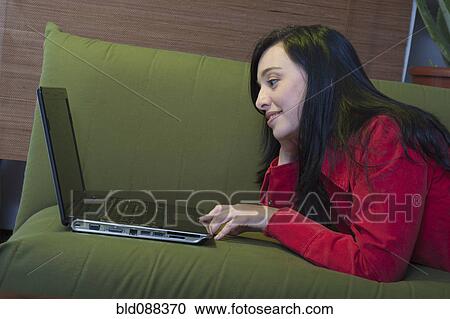 Stock Image - Hispanic woman laying on sofa using laptop. Fotosearch