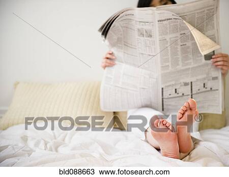 Stock Image - Korean woman reading newspaper in bed. Fotosearch