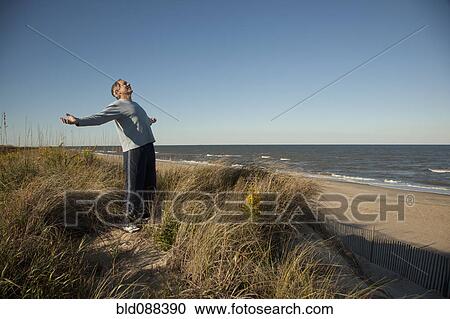 Stock Image - Man standing on sand dune near ocean with arms outstretched. Fotosearch