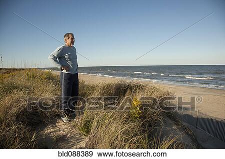 Stock Photo - Man standing on sand dune near ocean. Fotosearch