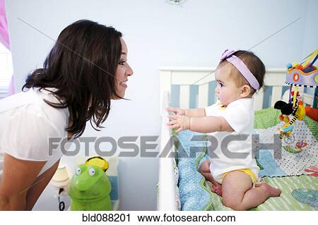 Stock Image - Mixed race mother looking at baby girl in crib. Fotosearch