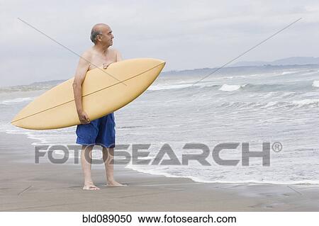 Stock Image - Senior Hispanic man holding surfboard on beach. Fotosearch