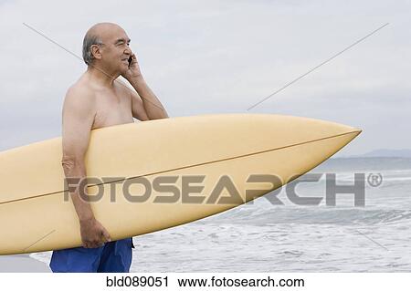 Stock Image - Senior Hispanic man holding surfboard on beach and talking on cell phone. Fotosearch