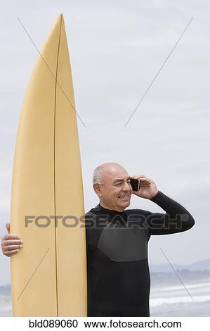 Stock Image - Senior Hispanic man holding surfboard on beach and talking on cell phone. Fotosearch