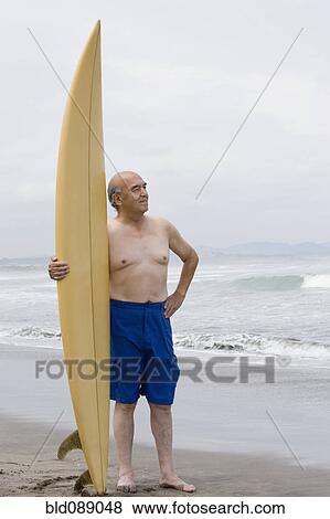 Stock Photo - Senior Hispanic man holding surfboard on beach. Fotosearch