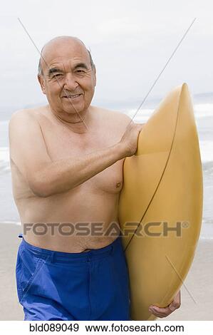 Stock Photo - Senior Hispanic man holding surfboard on beach. Fotosearch