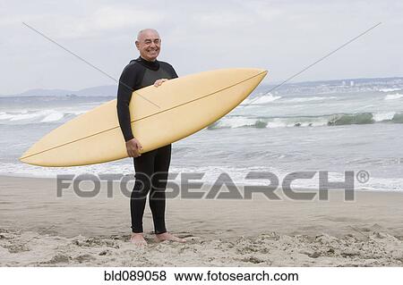 Stock Photo - Senior Hispanic man holding surfboard on beach. Fotosearch