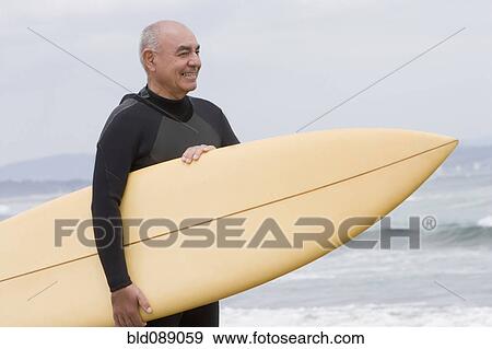 Stock Photo - Senior Hispanic man holding surfboard on beach. Fotosearch
