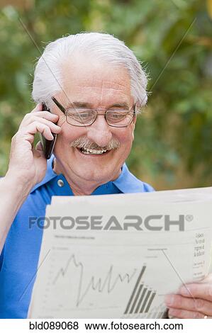 Stock Photo - Senior Hispanic man reading financial newspaper and talking on cell phone. Fotosearch