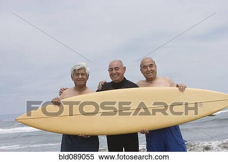 Stock Photography - Senior Hispanic men holding surfboard on beach. Fotosearch