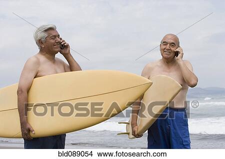 Picture - Senior Hispanic men holding surfboards on beach and talking on cell phones. Fotosearch
