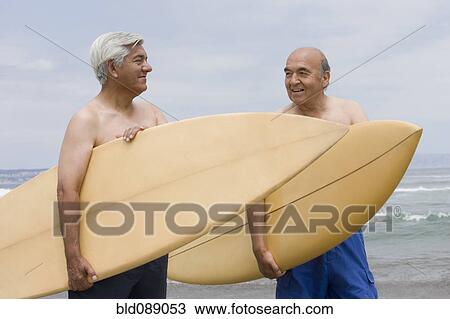 Stock Image - Senior Hispanic men holding surfboards on beach. Fotosearch