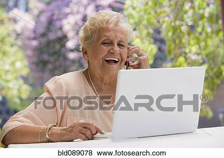 Stock Photo - Senior Hispanic woman using laptop and talking on cell phone. Fotosearch