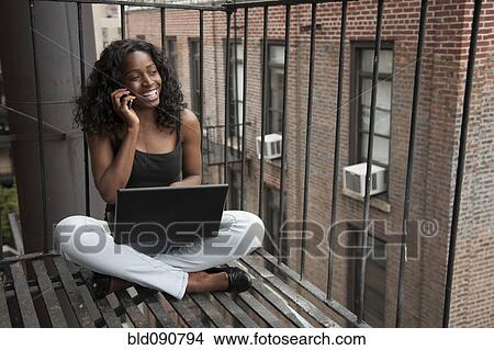 Picture - Black woman using laptop and cell phone on fire escape. Fotosearch