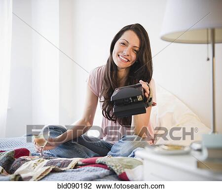 Stock Image - Brazilian woman sitting in bed with wine and instant camera. Fotosearch