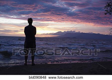 Stock Photo - Caucasian man standing on beach viewing sunset. Fotosearch