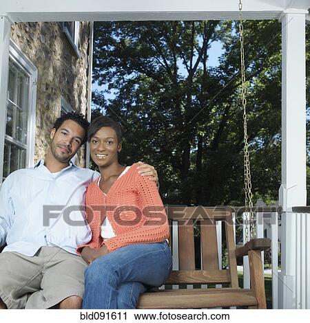 Stock Image - Mixed race couple sitting on porch swing. Fotosearch