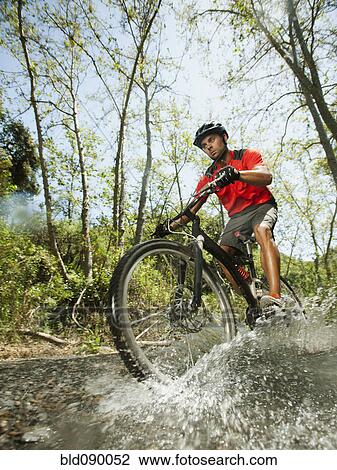 Stock Image - Mixed race man riding mountain bike through stream. Fotosearch