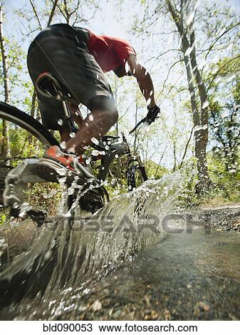 Stock Image - Mixed race man riding mountain bike through stream. Fotosearch