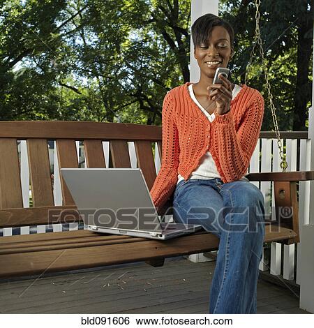 Stock Photograph - Mixed race woman on porch with cell phone and laptop. Fotosearch