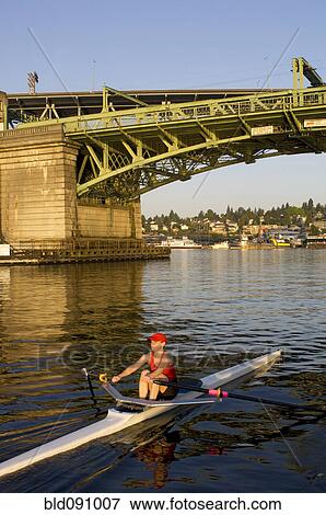 Stock Photo - Person rowing sculling boat on river under bridge. Fotosearch