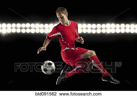 Stock Image - Soccer player jumping in mid-air kicking ball at night. Fotosearch