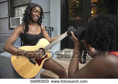 Stock Image - African American woman photographing sister playing guitar. Fotosearch