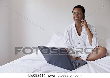Picture - African American woman using laptop and cell phone in bed. Fotosearch