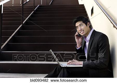 Stock Image - Asian businessman sitting on steps using cell phone and laptop. Fotosearch