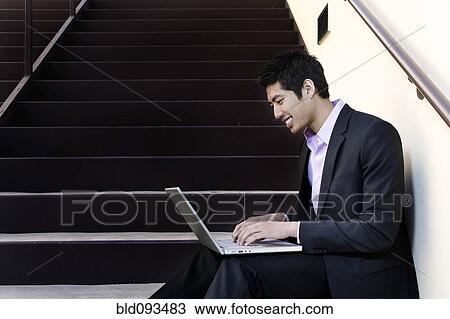 Stock Image - Asian businessman sitting on steps using laptop. Fotosearch