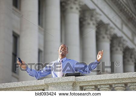Picture - Black businessman holding cell phone with arms outstretched. Fotosearch