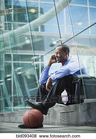 Stock Photo - Black businessman sitting outdoors with basketball talking on cell phone. Fotosearch
