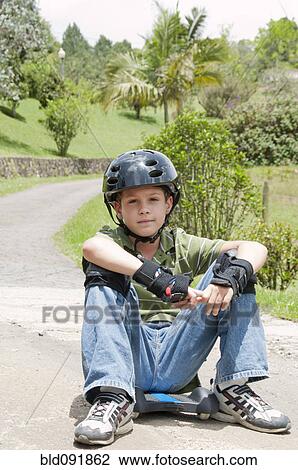 Stock Image - Hispanic boy in pads and helmet sitting on skateboard. Fotosearch