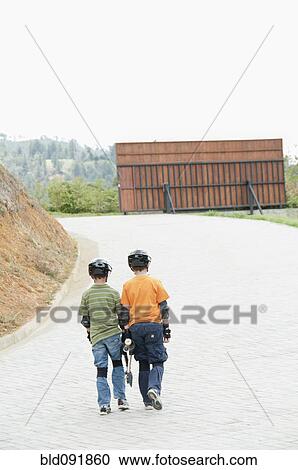Stock Image - Hispanic boys in pads and helmets carrying skateboard up road. Fotosearch