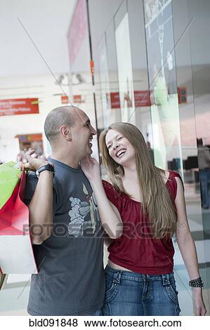 Stock Photo - Laughing Hispanic couple walking in mall carrying shopping bags. Fotosearch