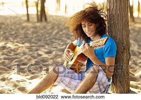 Stock Image - Mixed race teenager sitting in sand strumming guitar. Fotosearch