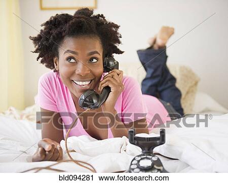 Stock Image - Black woman laying in bed talking on old-fashioned telephone. Fotosearch