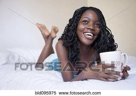Stock Photography - Black woman laying on bed drinking coffee. Fotosearch