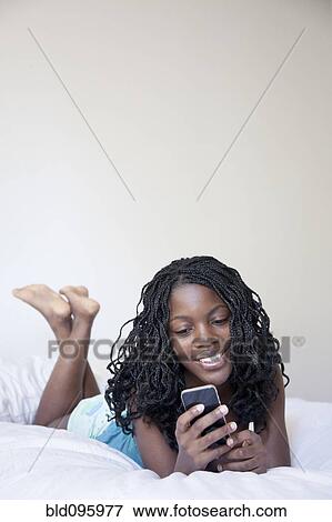 Stock Photo - Black woman laying on bed text messaging on cell phone. Fotosearch