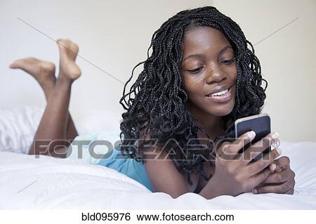 Stock Photograph - Black woman laying on bed text messaging on cell phone. Fotosearch