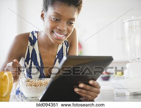 Stock Image - Black woman using digital tablet and eating cereal. Fotosearch
